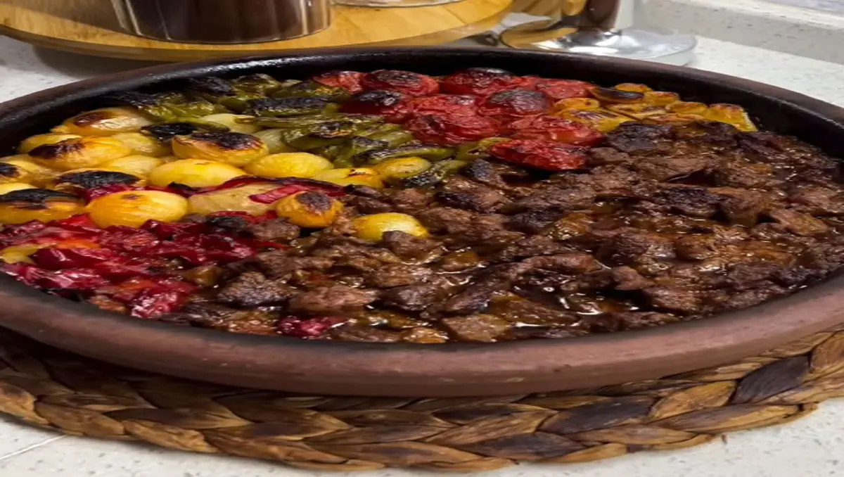 Kahverengi topraktan yapılmış oval bir güveç kabında fırında pişmiş et ve sebzelerin yakın çekim görüntüsü. Kabın sağ tarafında kahverengi soslu, küçük kuşbaşı doğranmış et parçaları yer alıyor. Sol tarafta, üstten alta doğru sıra sıra dizilmiş kavrulmuş sebzeler bulunuyor: En üstte yeşil biberler, ortada küçük sarı patatesler ve en altta kırmızı biber veya domates parçaları var. Sebzelerin bazı kısımları hafifçe kararmış, bu da fırında güzelce kızardıklarını gösteriyor. Yemek, kahverengi örgülü bir hasır servis altlığının üzerinde duruyor. Arka planda beyaz bir tezgah ve bulanık mutfak eşyaları görülüyor. Yemek buharı üzerinde ve taze pişmiş görünüyor, iştah açıcı bir sunum sergiliyor.