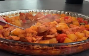 Buharı tüten, cam fırın kabında servis edilmeye hazır, fırınlanmış tavuk ve patates yemeği. Yemeğin içinde kızarmış tavuk parçaları, küp doğranmış sarı patatesler, kırmızı ve yeşil biberler görülüyor. Bir el metal bir servis kaşığı ile yemeği karıştırıyor veya servis yapıyor. Yemek sıcak ve taze görünüyor, kahverengi ve altın sarısı tonları hakim.
