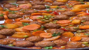 Yakın çekim tencere içinde pişmiş, buharı tüten, bol soslu ve renkli bir sebzeli köfte yemeği. Kahverengi, yuvarlak köfteler; parlak turuncu halka havuçlar, sarı küp patatesler ve canlı yeşil bezelyeler kırmızı-kahverengi koyu domates bazlı sosun içinde dağılmış durumda. Üzerine taze kıyılmış maydanoz serpilmiş. Yemek, ev yapımı, besleyici ve sıcak bir lezzet vaat eden çekici bir görünüme sahip. Görüntü, iştah açıcı ve rahatlatıcı bir atmosfer sunuyor.