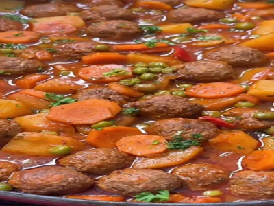 Yakın çekim tencere içinde pişmiş, buharı tüten, bol soslu ve renkli bir sebzeli köfte yemeği. Kahverengi, yuvarlak köfteler; parlak turuncu halka havuçlar, sarı küp patatesler ve canlı yeşil bezelyeler kırmızı-kahverengi koyu domates bazlı sosun içinde dağılmış durumda. Üzerine taze kıyılmış maydanoz serpilmiş. Yemek, ev yapımı, besleyici ve sıcak bir lezzet vaat eden çekici bir görünüme sahip. Görüntü, iştah açıcı ve rahatlatıcı bir atmosfer sunuyor.